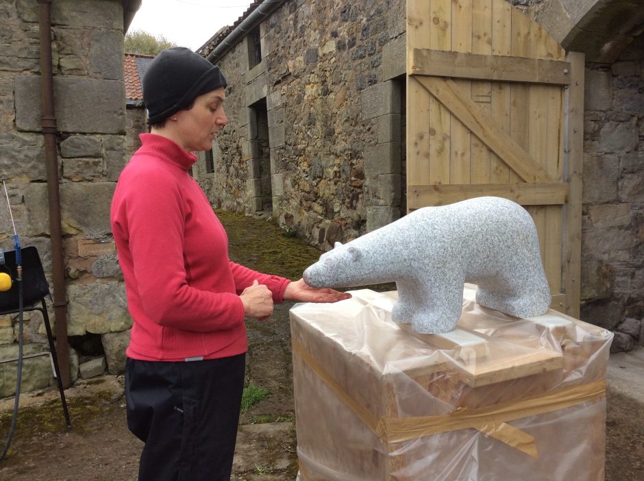 Marion Smith wearing bright pink top with pale grey granite carving of a polar bear. The carving sits on top of a big wooden box covered in polythene, there is an open barn door behind, stone built buildings in background.