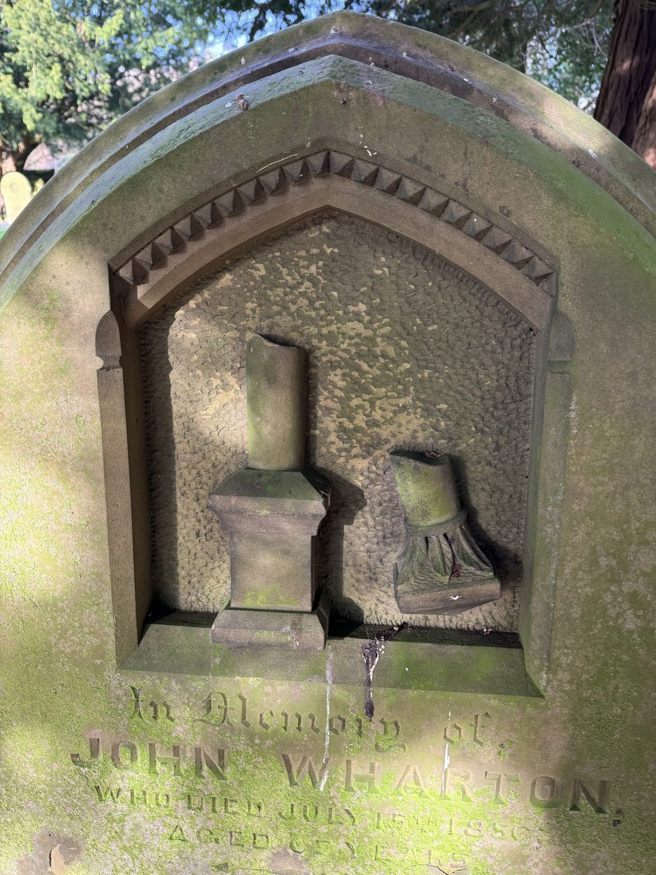Detailed image of the top of a sandstone headstone, it has the detail of a broken column in a recess with a decorated, arched top. It is inscribed 'Im Memory of John Wharton'.