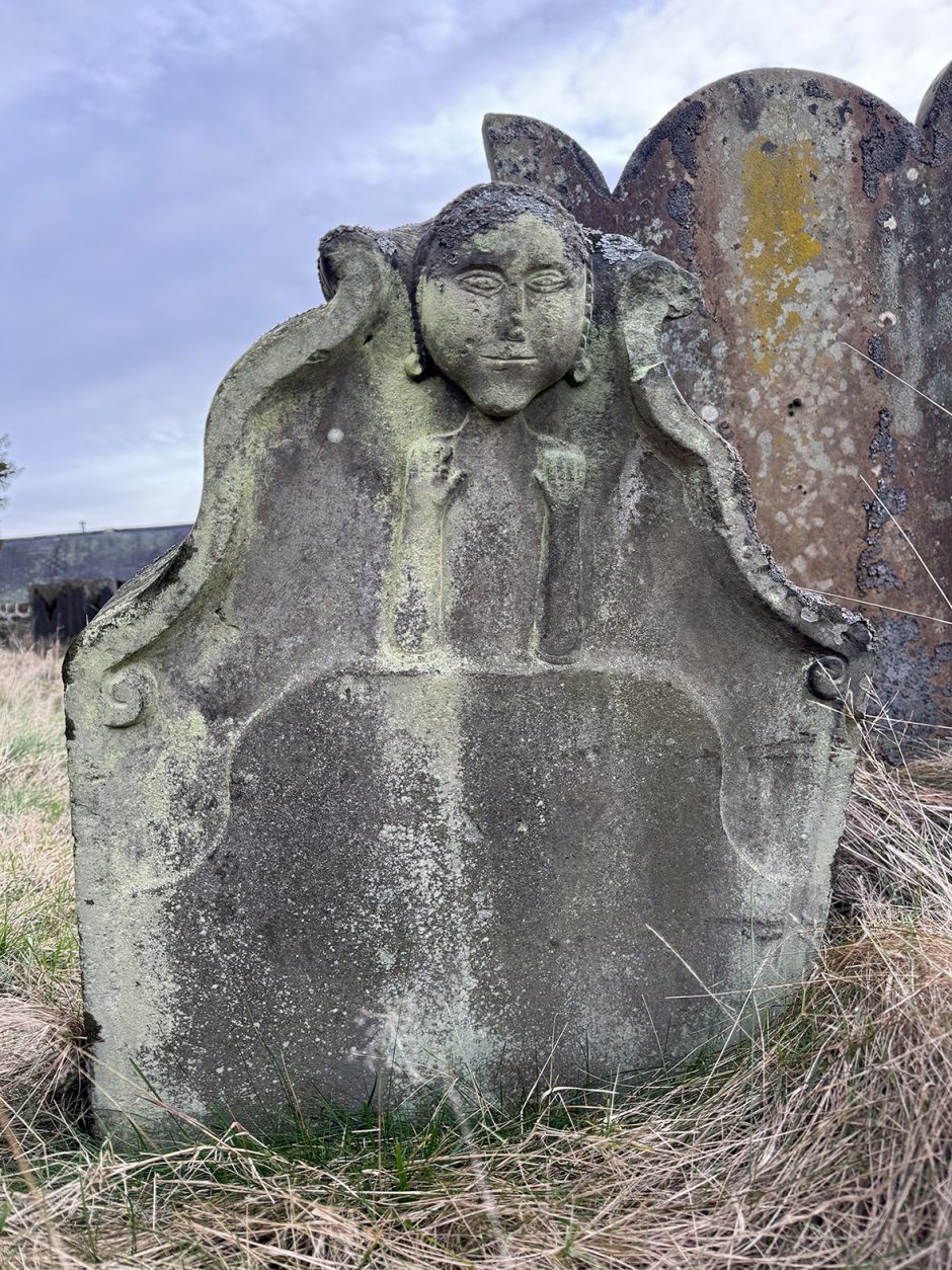 Odd shaped sandstone headstone with a girl's head at the top, her arms are up, with her hands at shoulder height, one of the palms faces outwards, the other inwards. In the background on the right is the unusual outline of another headstone.