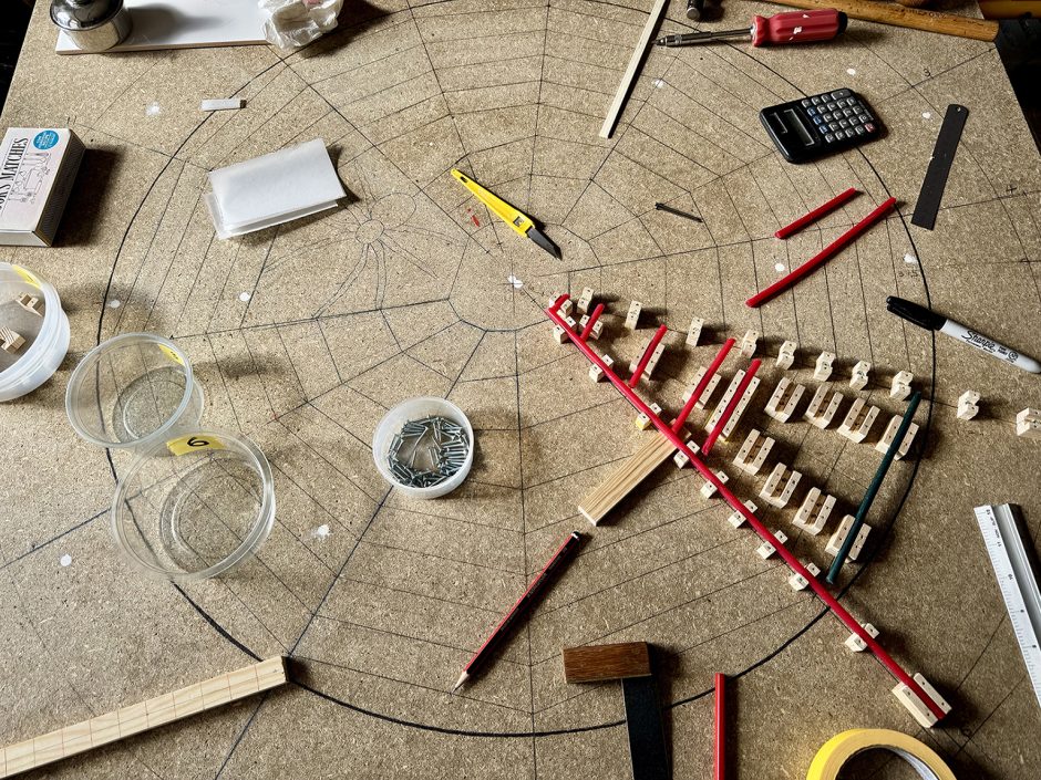 Tools and materials lying on top of a drawing of a spider's web. Tools include from top right a hammer, screwdriver, calculator and ruler. There are some red strips of wax in a jig made of wood
